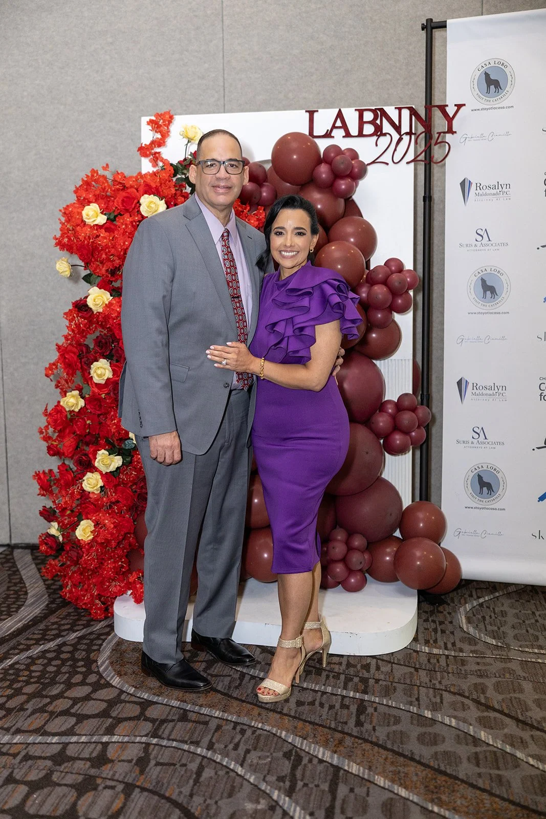 A man and woman standing together at a formal event with a backdrop decorated with balloons and flowers. The man is wearing a gray suit with a maroon patterned tie, glasses, and black shoes. The woman is dressed in a purple dress with ruffled sleeves