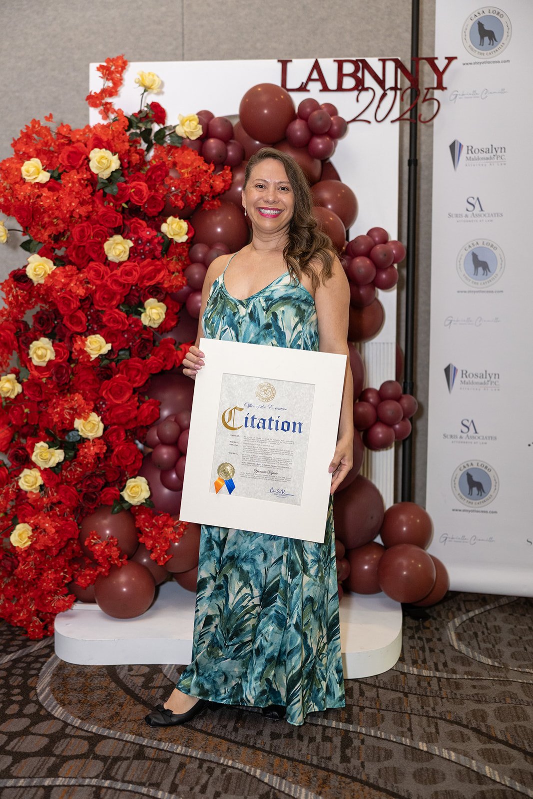 A woman in a long green and blue floral dress holding a framed certificate at an event with a red and yellow floral arrangement and a balloon display in the background, celebrating Lanny 2025.