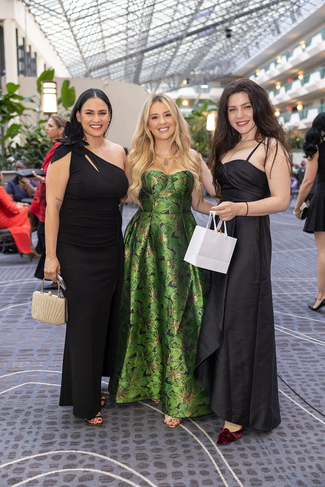 Three women in formal dresses stand together, smiling at an indoor event, with plants and other guests in the background.