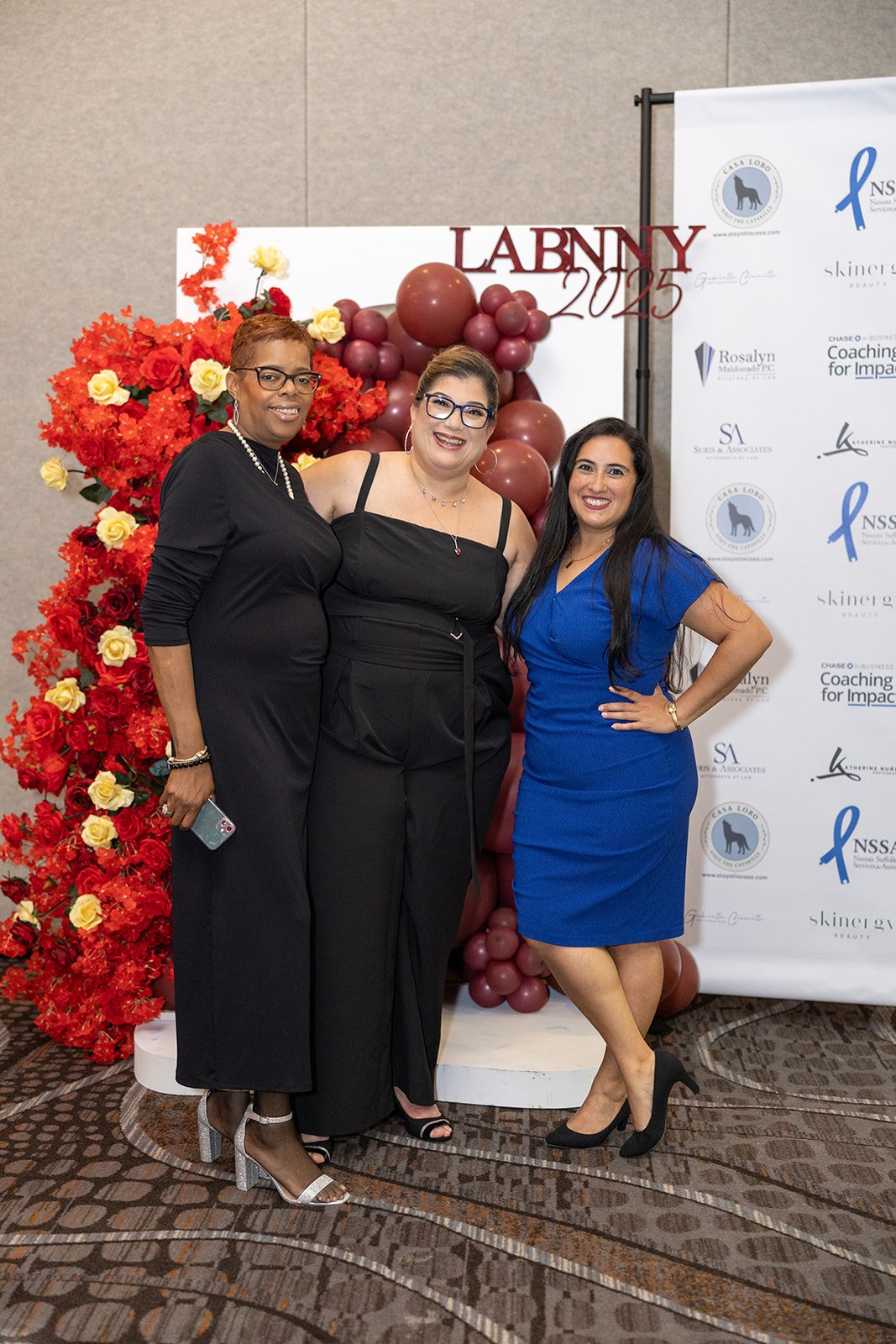 Three women in formal attire standing together in front of a red floral display and a backdrop with event logos. One woman is wearing a black dress, another a black top and pants, and the third a blue dress.