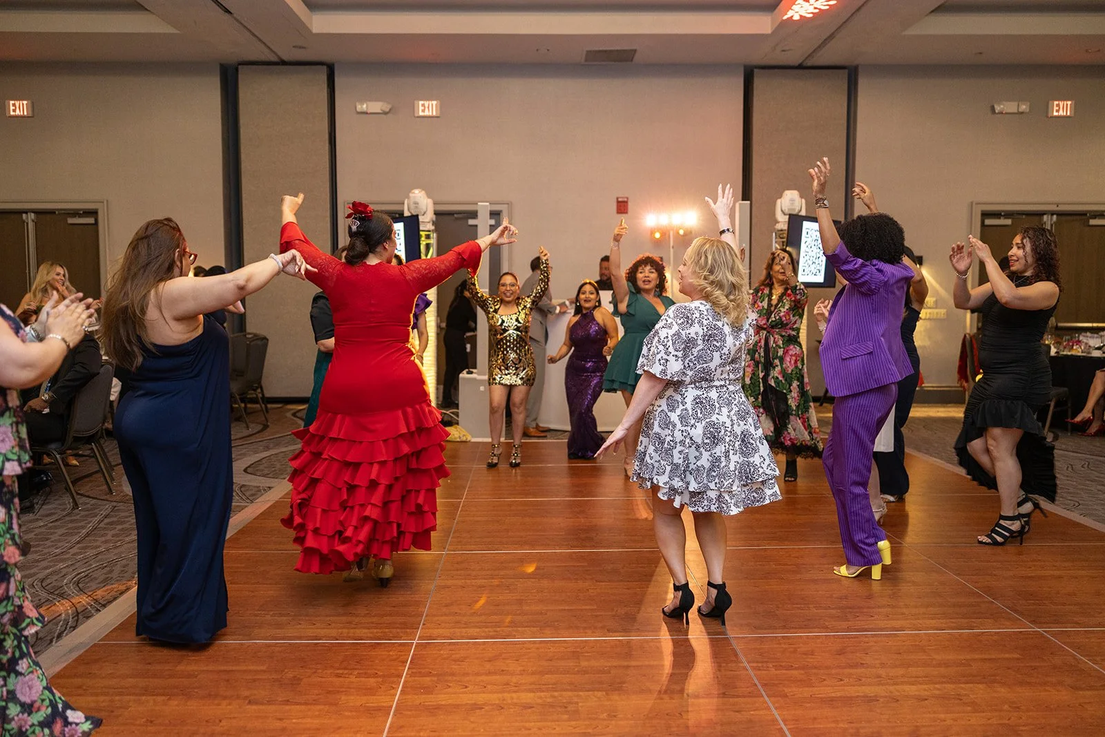 People dancing and celebrating at an indoor event with a wooden floor, some women wearing colorful dresses, and a festive atmosphere.