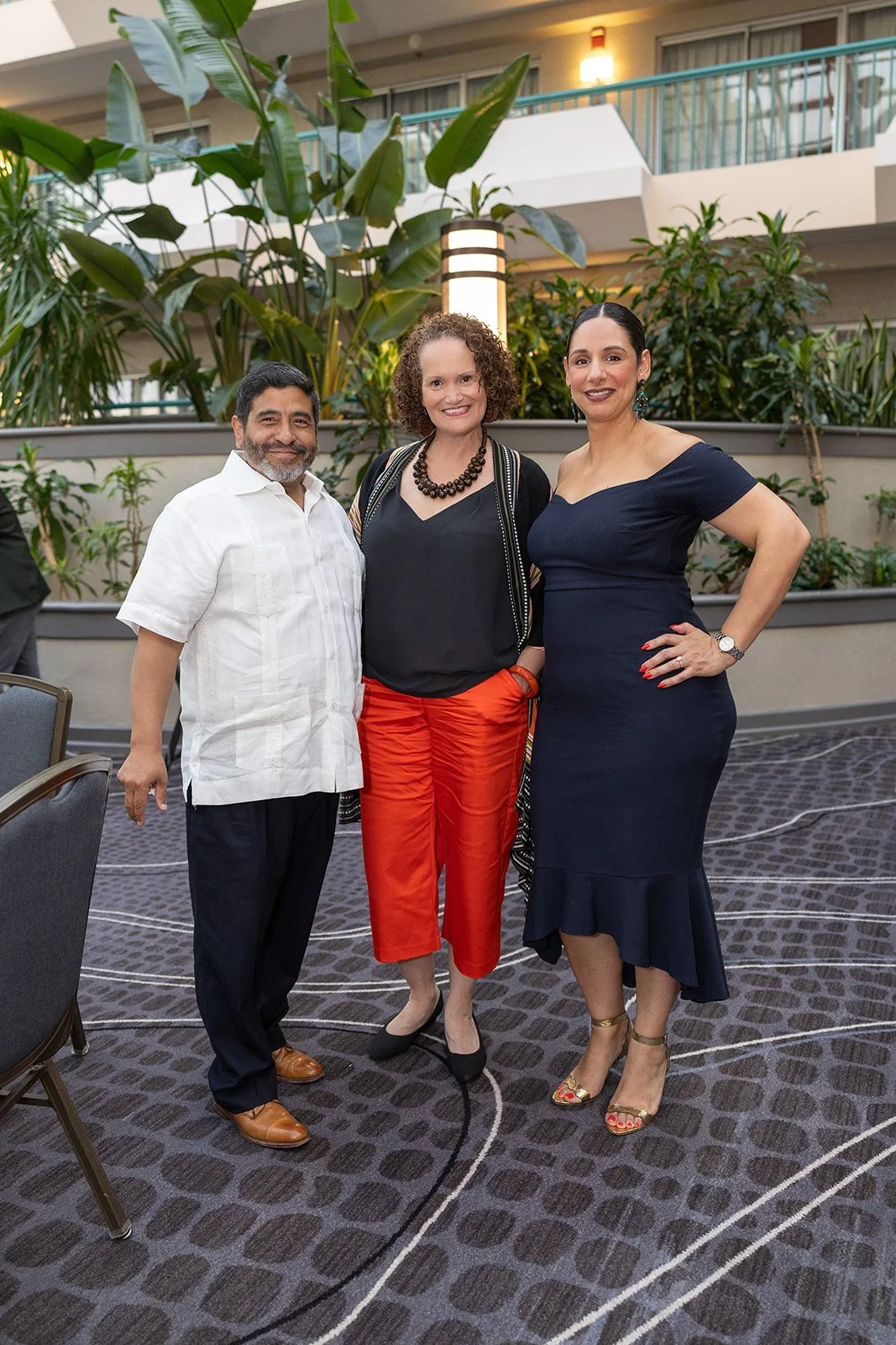Three people standing together indoors, a man and two women, smiling for the camera, with plants and a balcony in the background.