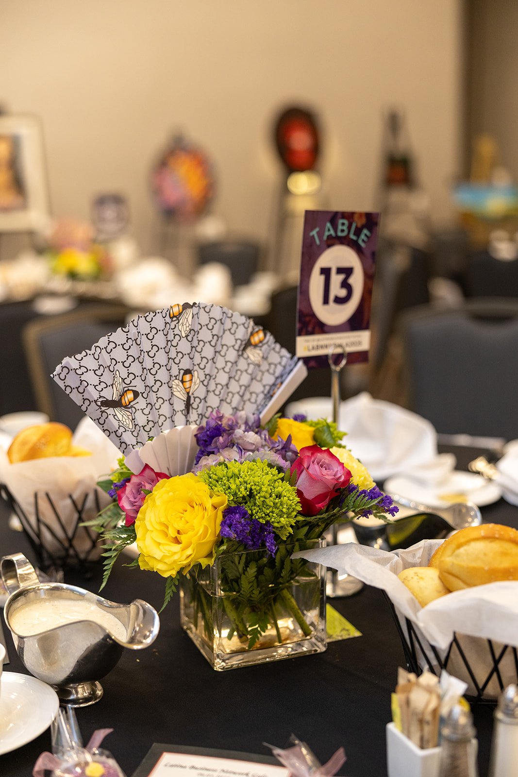 A table centerpiece with a colorful flower arrangement in a glass vase, featuring yellow, pink, purple, and green flowers, along with a table number 13 sign and bee-themed napkins. The table is set for a formal event with plates, cutlery, and bread b