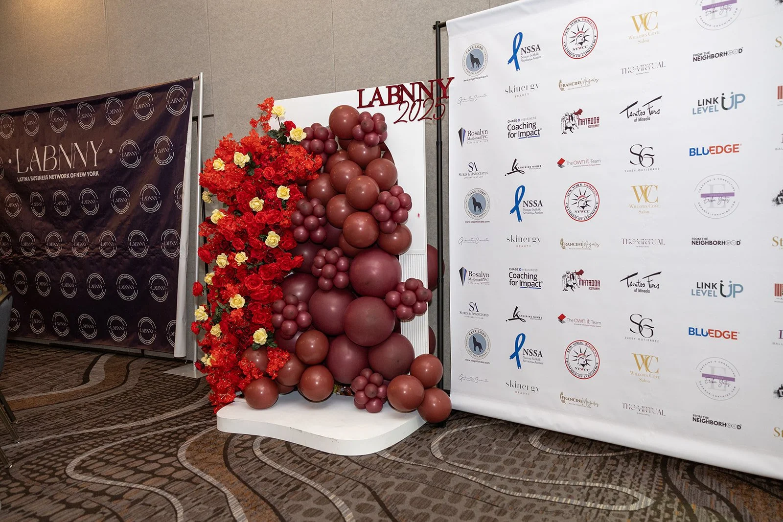 Decorative display of red and burgundy balloons and red and yellow flowers next to a backdrop with event logos and text that reads "LABNY 2023"
