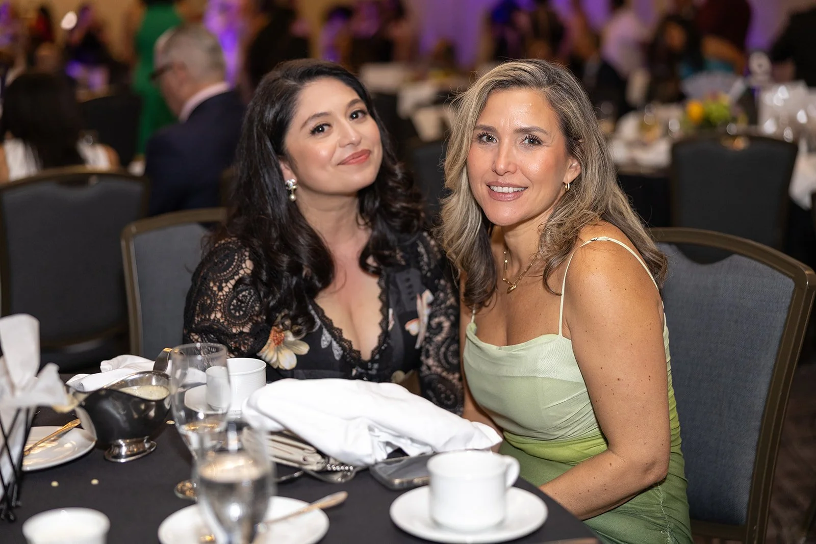 Two women sitting at a banquet table smiling at the camera during a formal event.