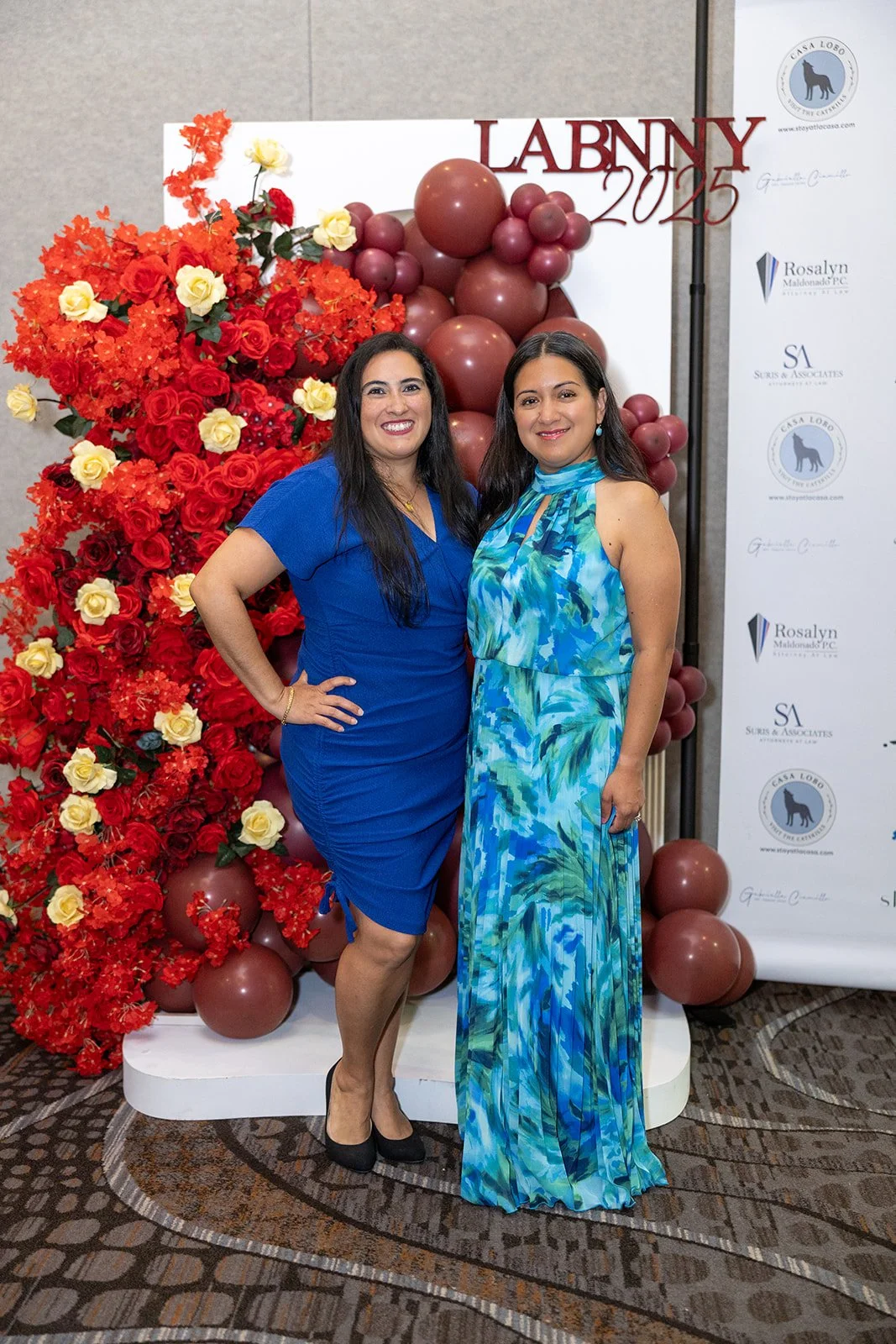 Two women dressed in blue and teal dresses standing in front of a floral and balloon backdrop at a graduation event for LabNNY 2023.
