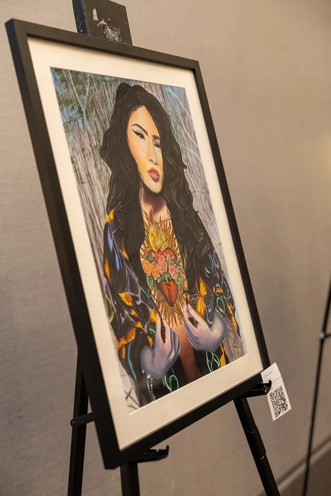 A framed colorful artwork of a woman with long dark hair, holding her hands over her chest with a tattoo of a heart surrounded by flowers on her chest. The background features a natural setting with trees.