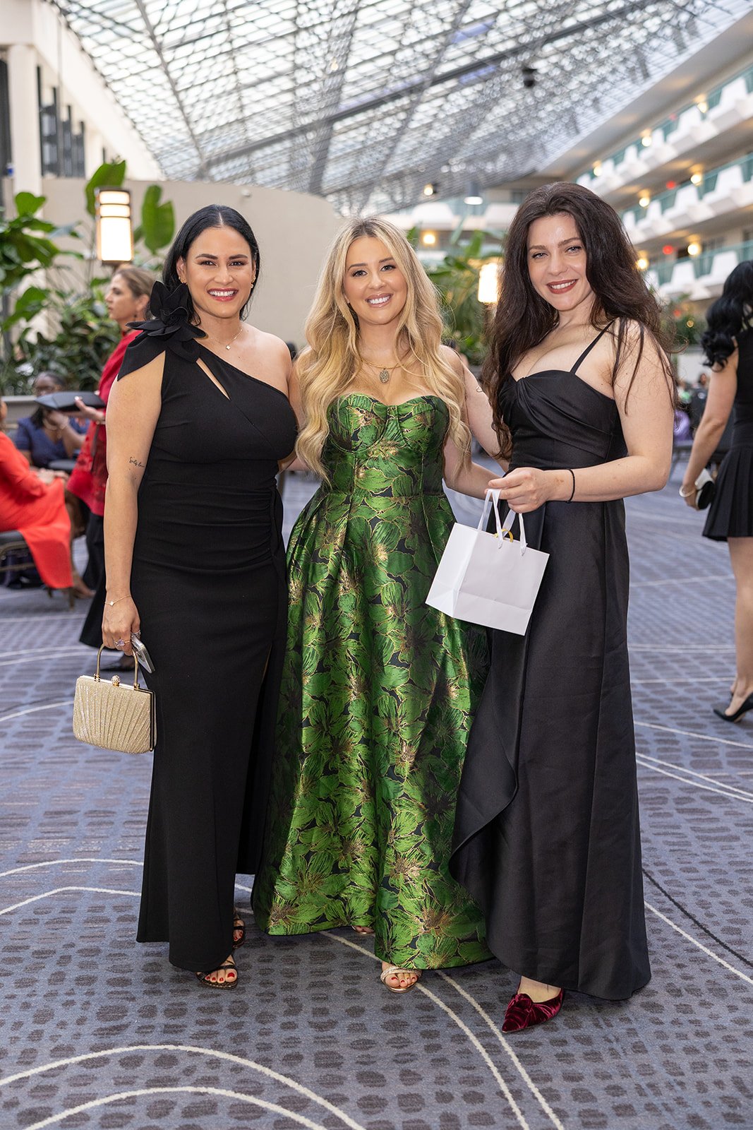Three women in evening dresses standing together at an event, smiling at the camera, inside a modern building with a glass ceiling and plants.