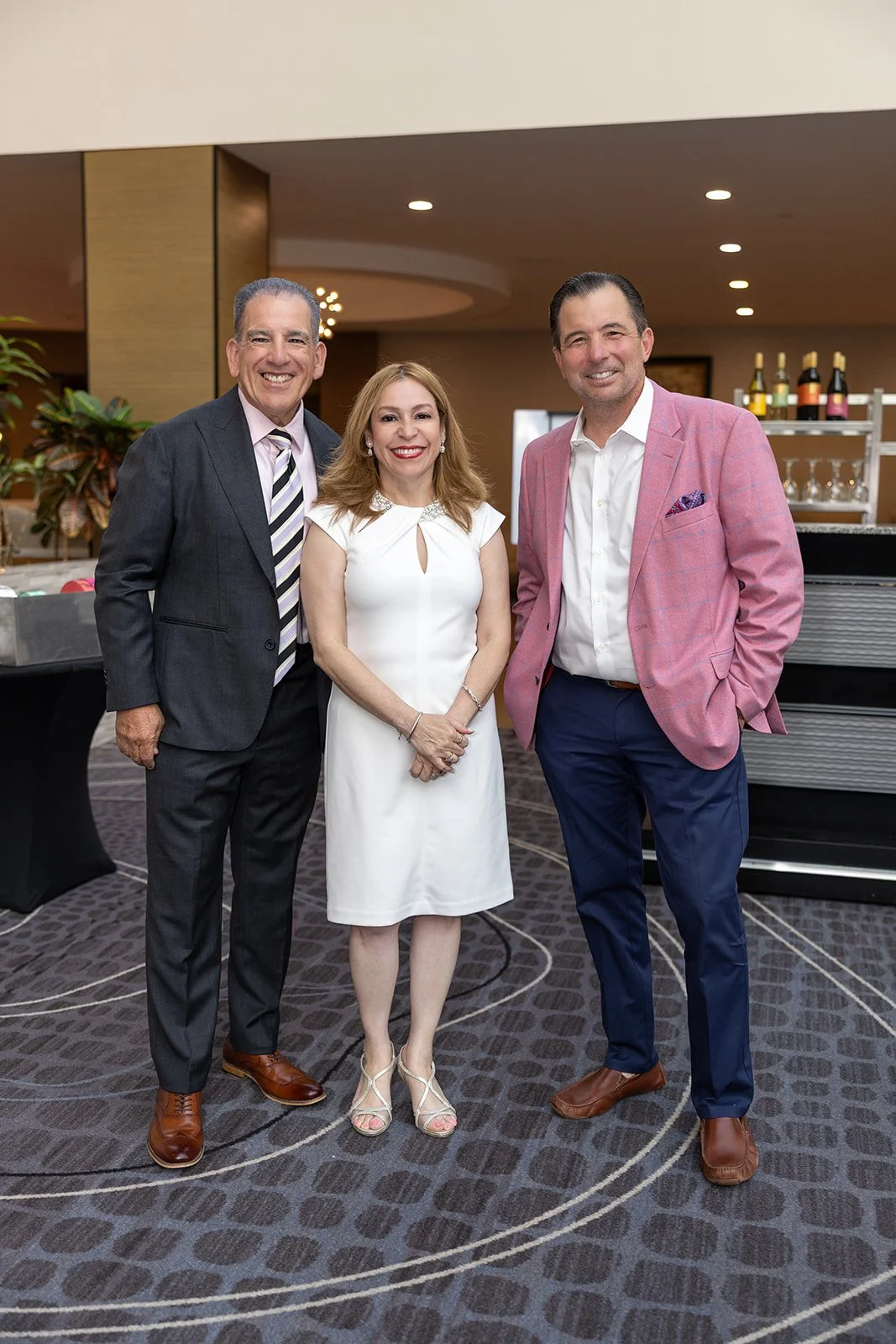 Three people in formal attire standing together at an indoor event, smiling at the camera.
