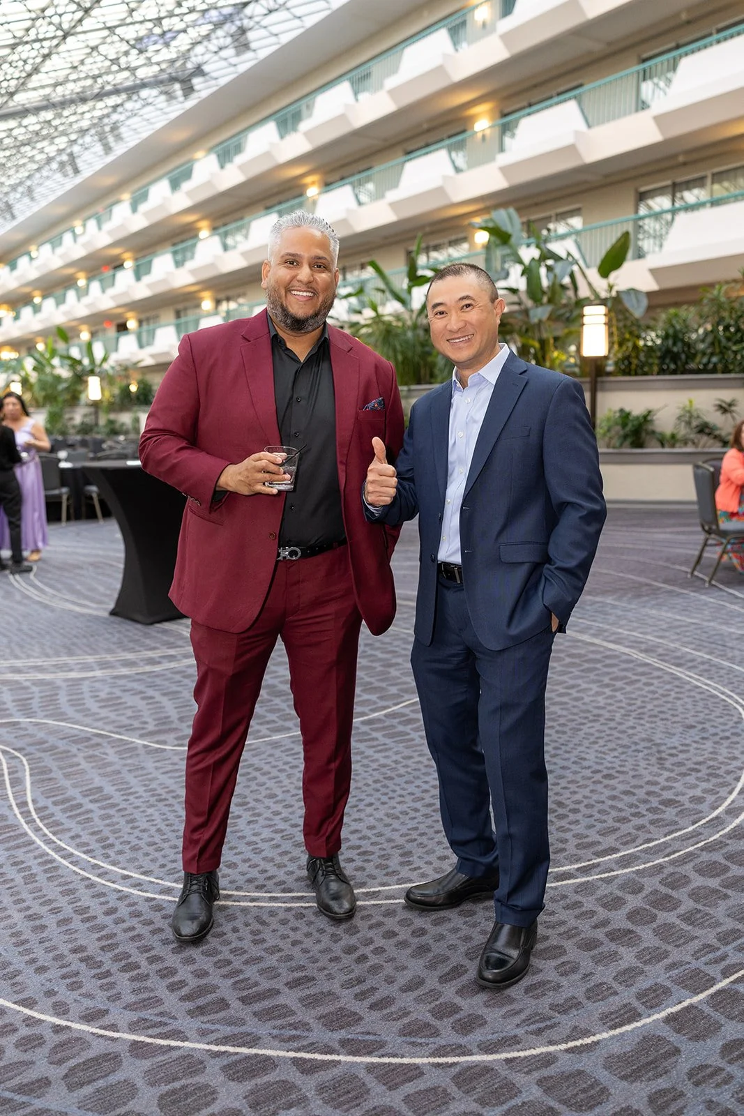 Two men in suits standing in a spacious indoor atrium, smiling and posing for a photo at a formal event. The man on the left wears a burgundy suit and holds a drink, while the man on the right wears a navy suit and gives a thumbs-up. There are other 