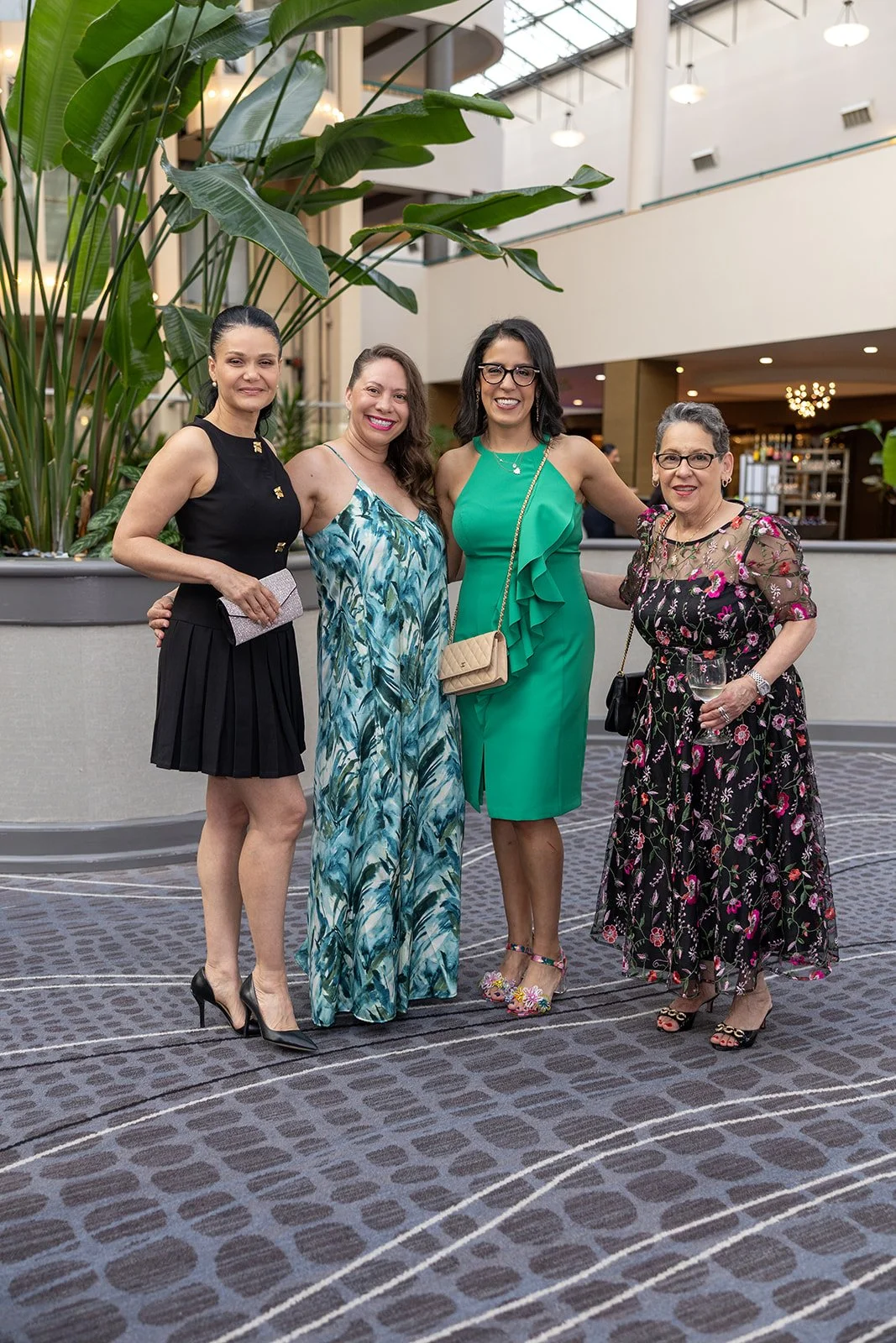 Four smiling women in stylish dresses and heels standing together inside a modern building with indoor plants and high ceilings.
