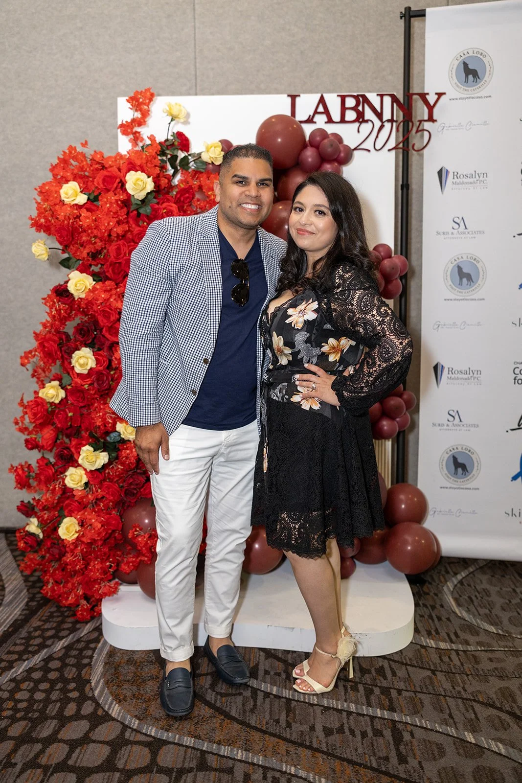 A man and woman posing together at a formal event with a red floral backdrop and balloons, standing on a patterned carpet.