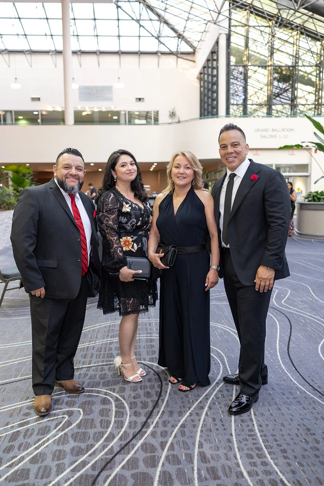 Four people dressed in formal and semi-formal attire standing inside a large, modern atrium with glass ceiling and plants.