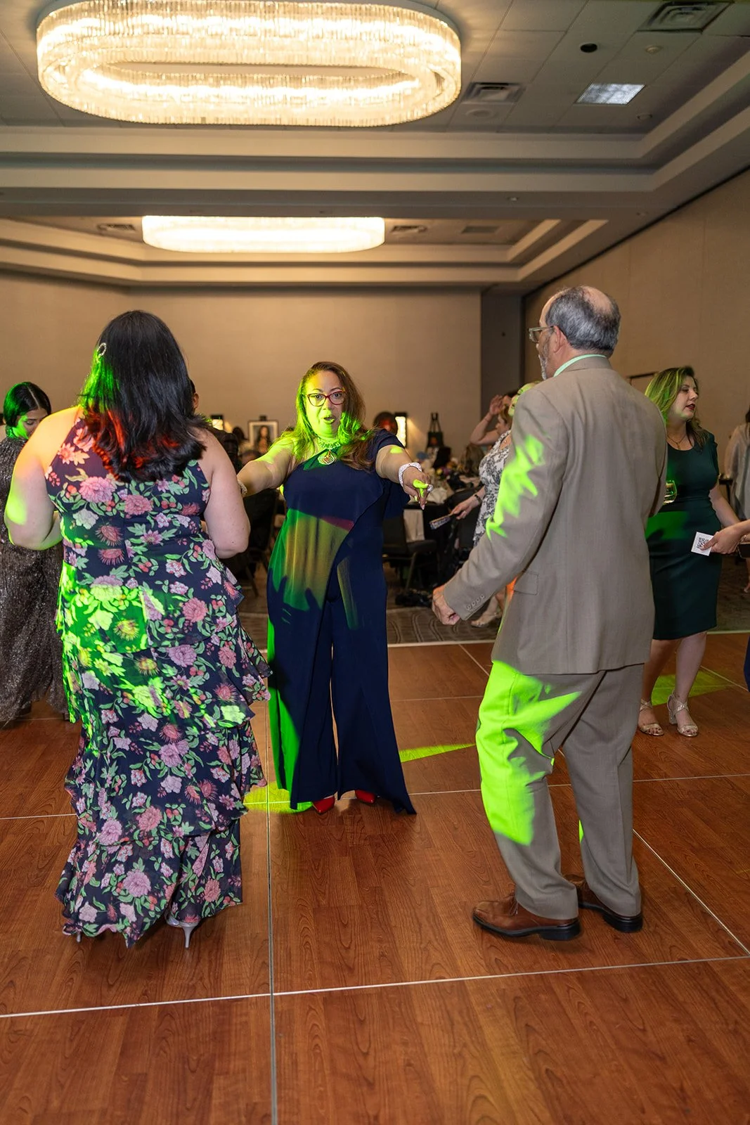 People dancing at a formal event or wedding reception in a ballroom with a high ceiling and large chandelier.