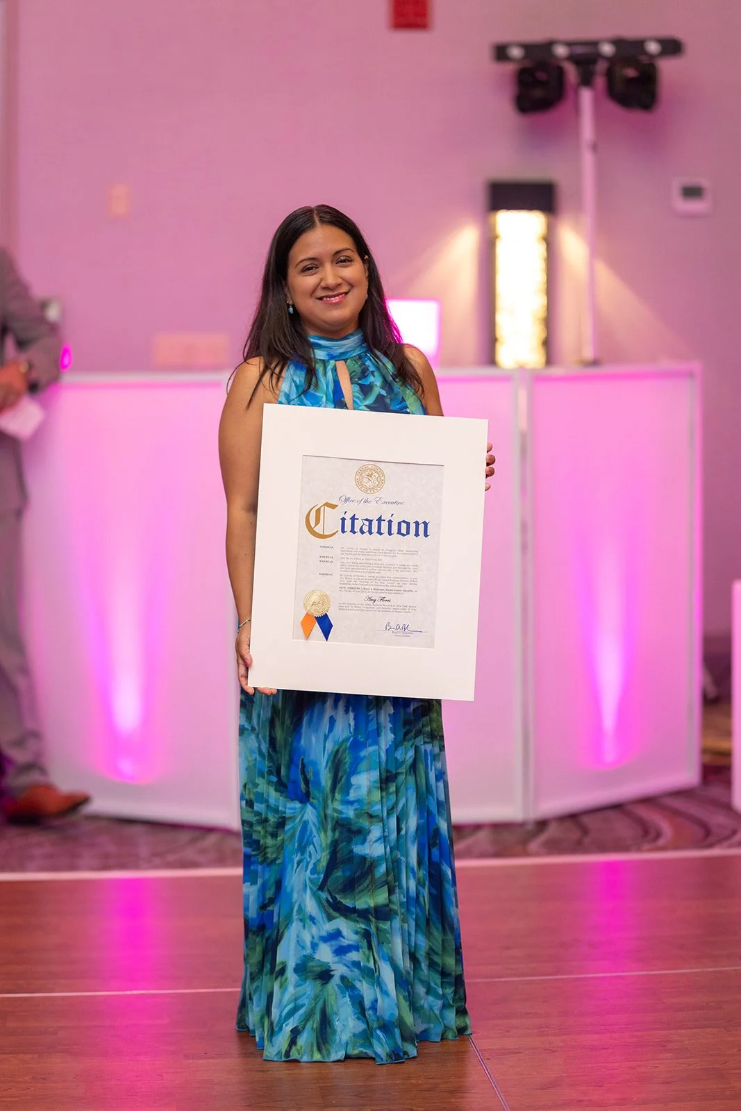 A woman in a blue and green dress holding a framed certificate at an indoor event with purple lighting and a DJ booth in the background.