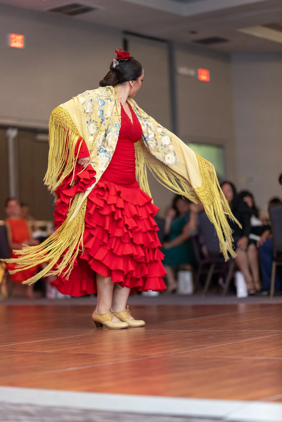 A woman in a red flamenco dress with ruffles and a yellow shawl with floral embroidery and long fringe, dancing on a wooden floor at an indoor event.