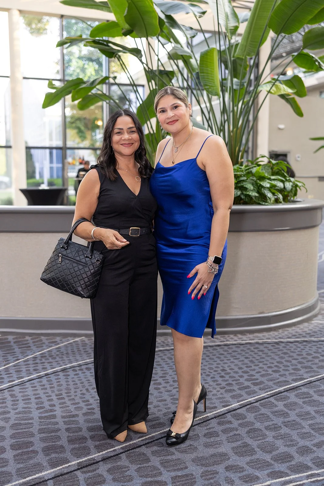 Two women standing together indoors, smiling, with large green plants in the background, at what appears to be a social or formal event.
