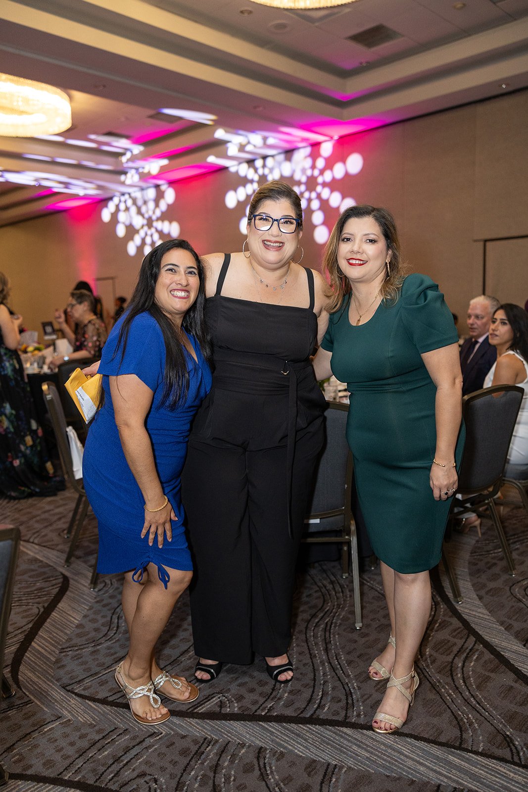 Three women standing together at an indoor event, smiling, with people seated at tables in the background and pink and white lighting decorations on the ceiling.