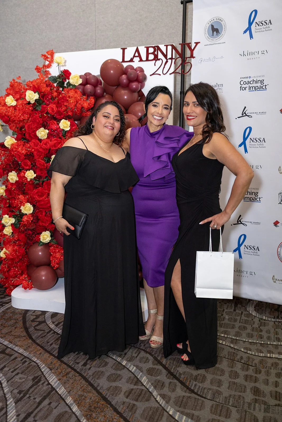 Three women dressed in formal attire posing together at an event, with a backdrop and floral decorations.