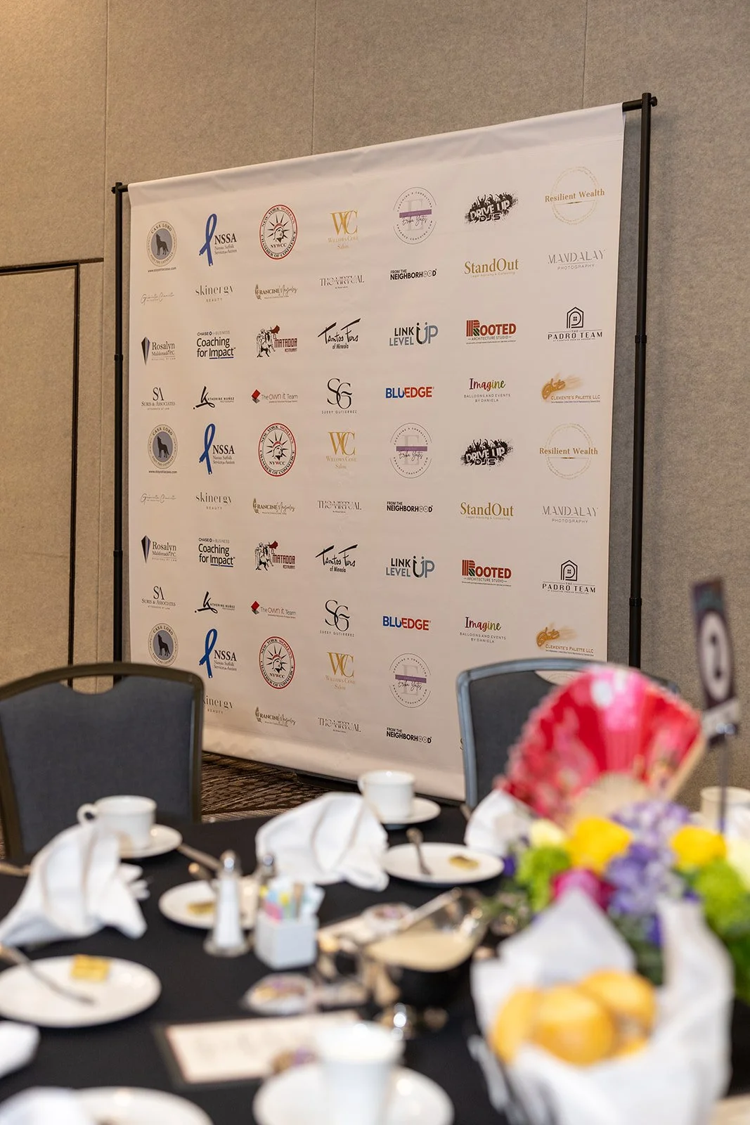 Event backdrop with logos, a round table with white cups, plates, and a floral centerpiece, in a conference or banquet room.