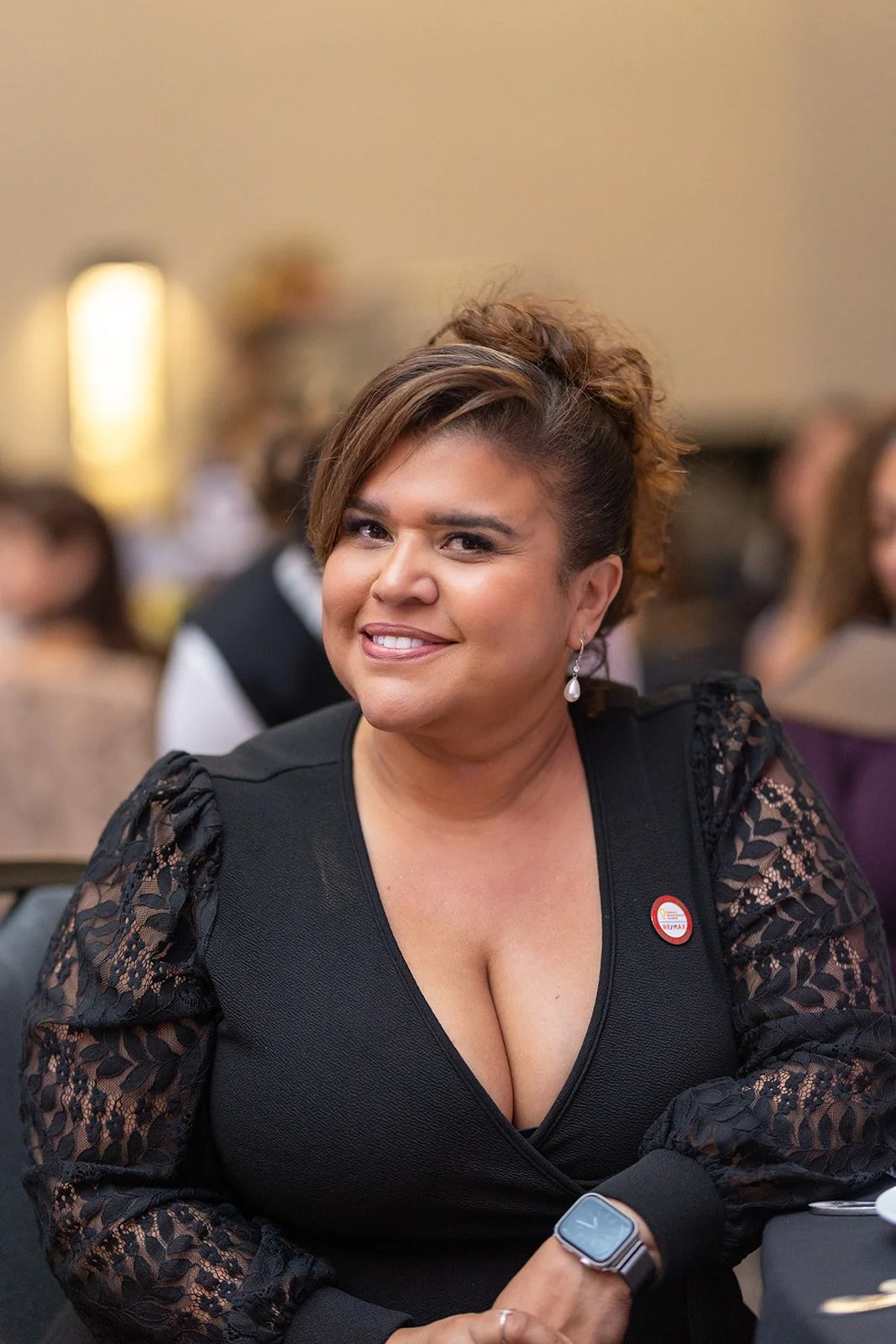 A woman with short brown hair in an elegant black dress with lace sleeves, smiling and sitting at a table during an event.