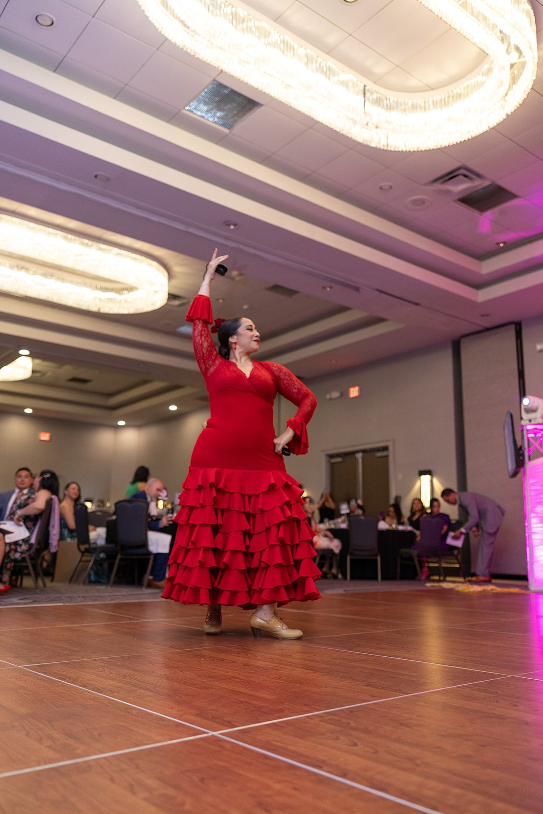 A woman in a red flamenco dress with ruffled skirt performs on a wooden dance floor at an indoor event, with an audience seated at tables in the background.