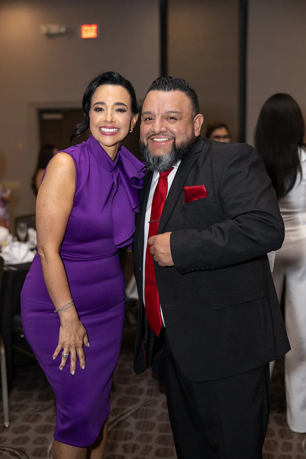 A woman in a purple dress smiling and standing next to a man in a black suit with a red tie, both at a formal event.