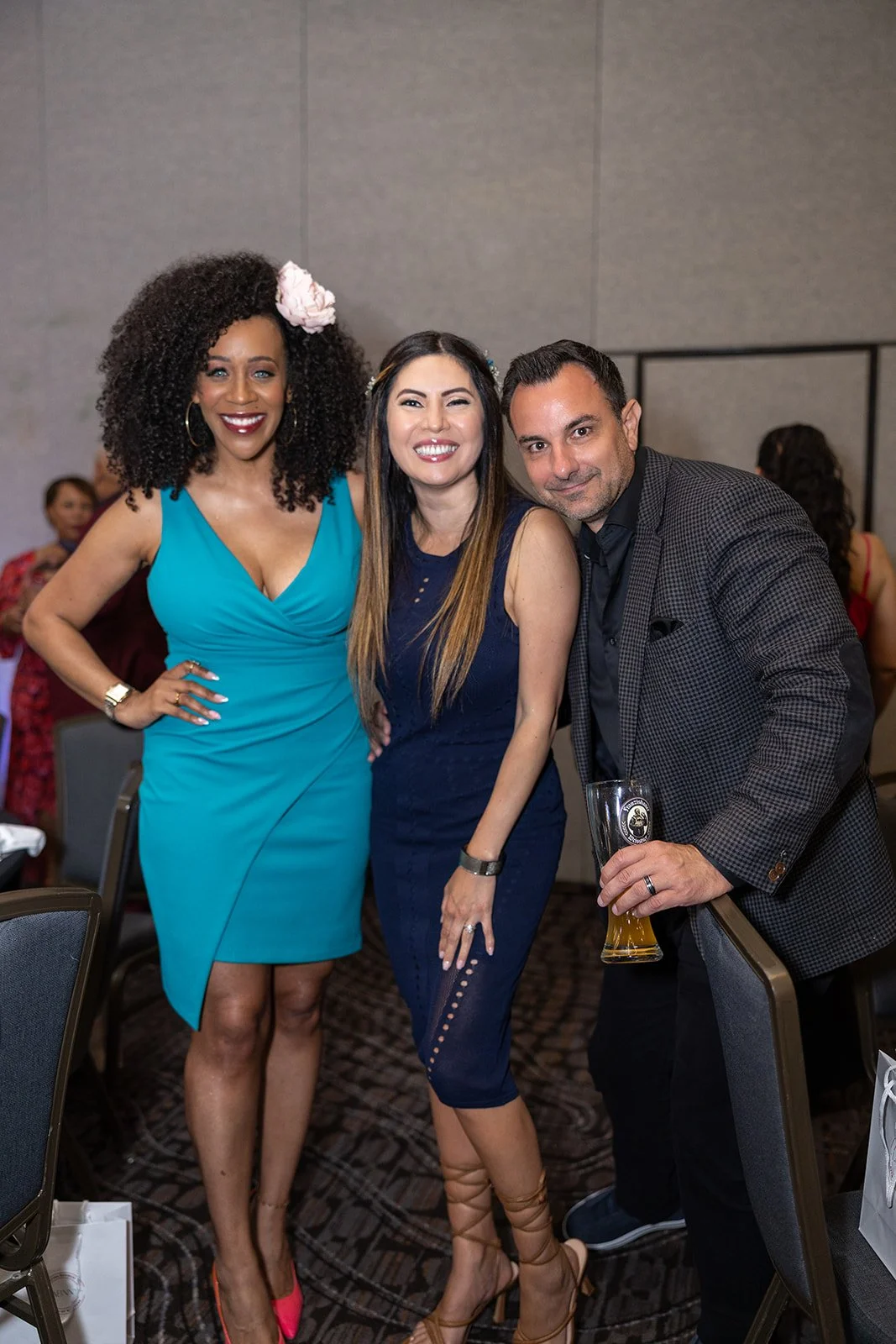 Three people at a social event posing for a photo, two women and one man. The woman on the left is wearing a bright blue dress with pink shoes, the woman in the middle is in a navy blue dress, and the man on the right is in a checkered blazer holding