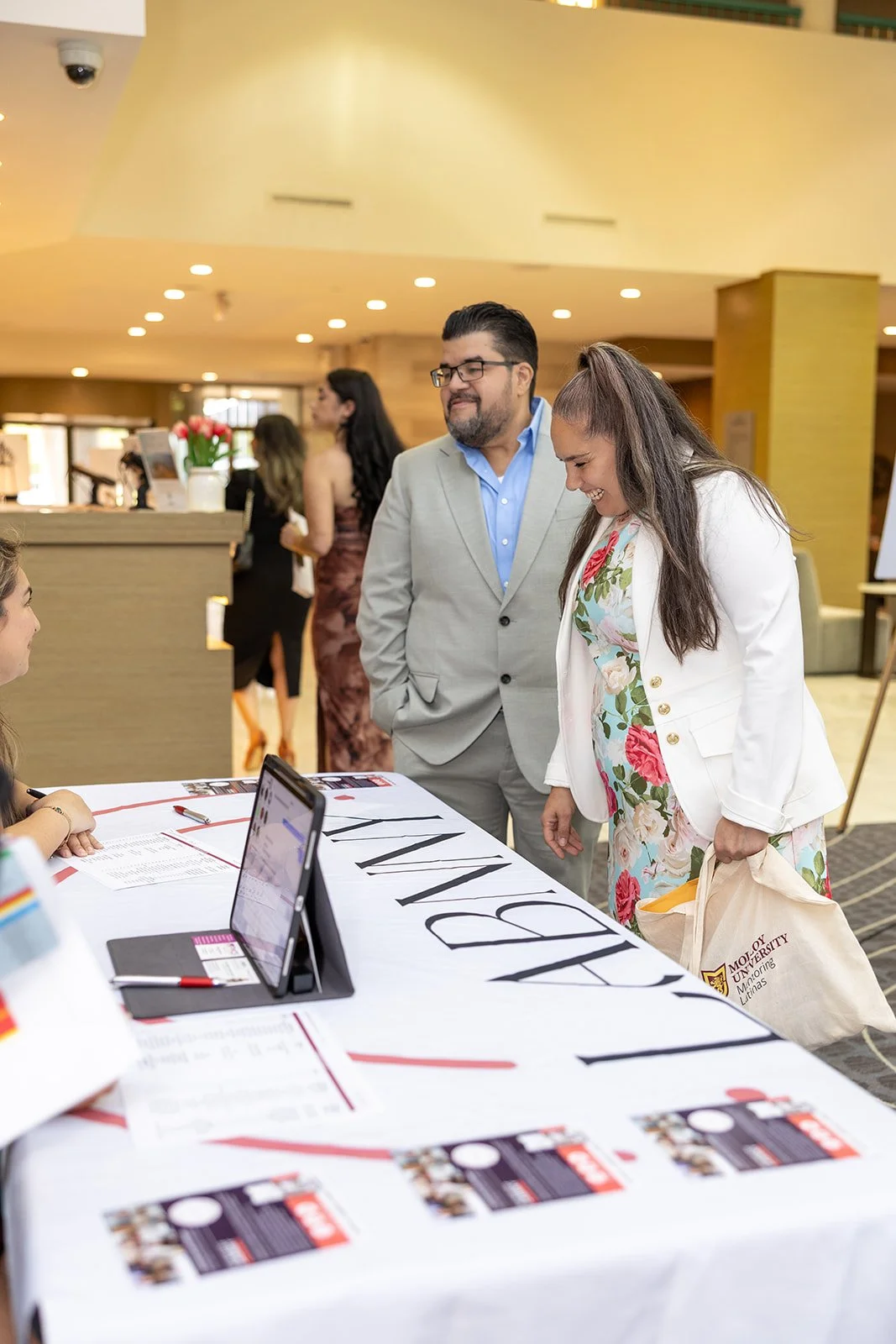 Two people, a man and a woman, talking to a staff member at a table with a sign reading 'Library'. The woman is holding a tote bag from the University of Minnesota. The setting is a busy indoor event space.