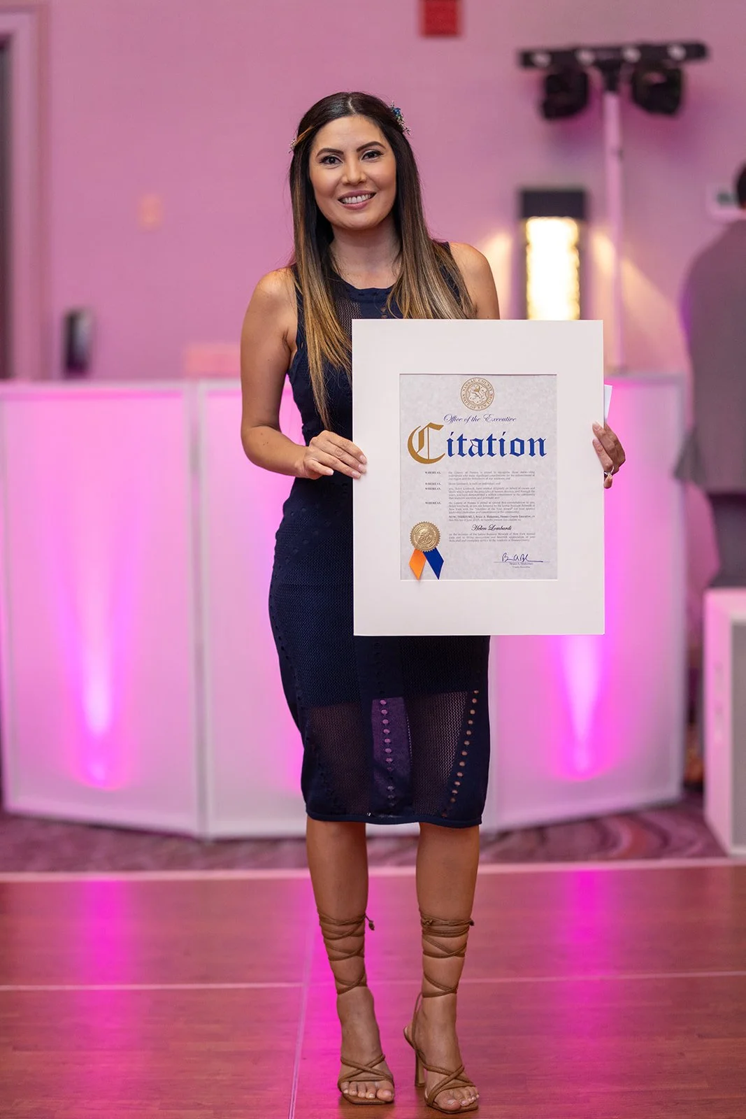 A woman in a black sleeveless dress holding a large framed certificate at an event with pink lighting.