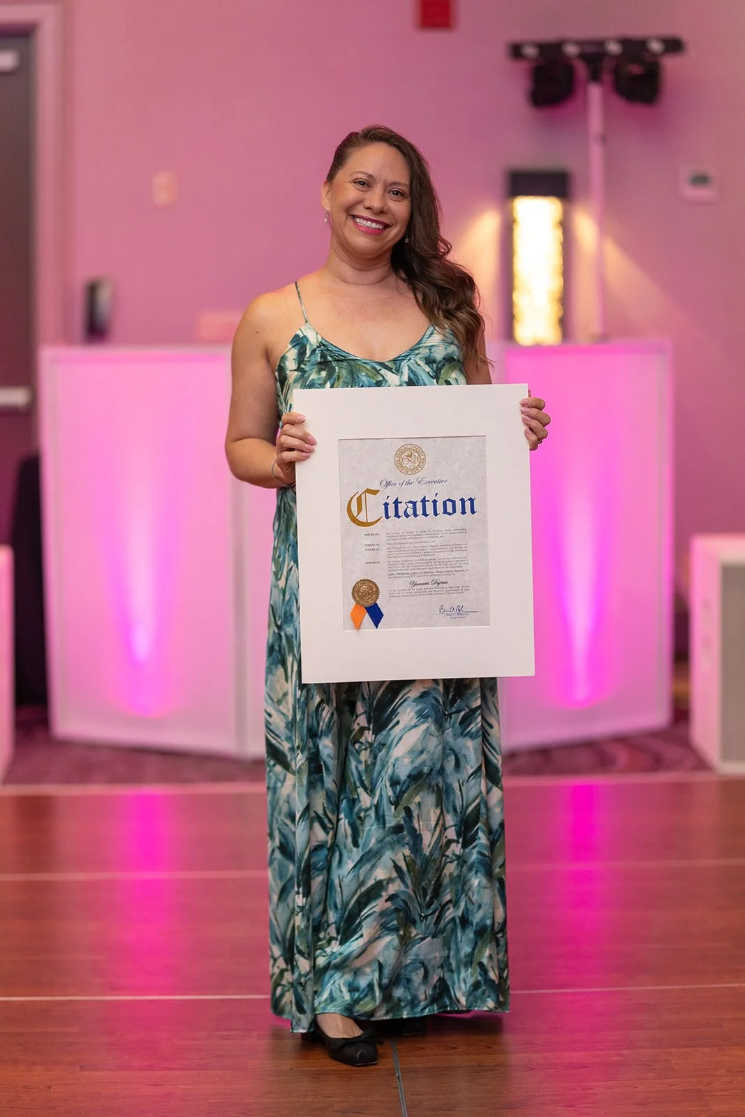 Woman in a long, patterned dress smiling and holding a framed certificate or award at an indoor event with pink lighting.
