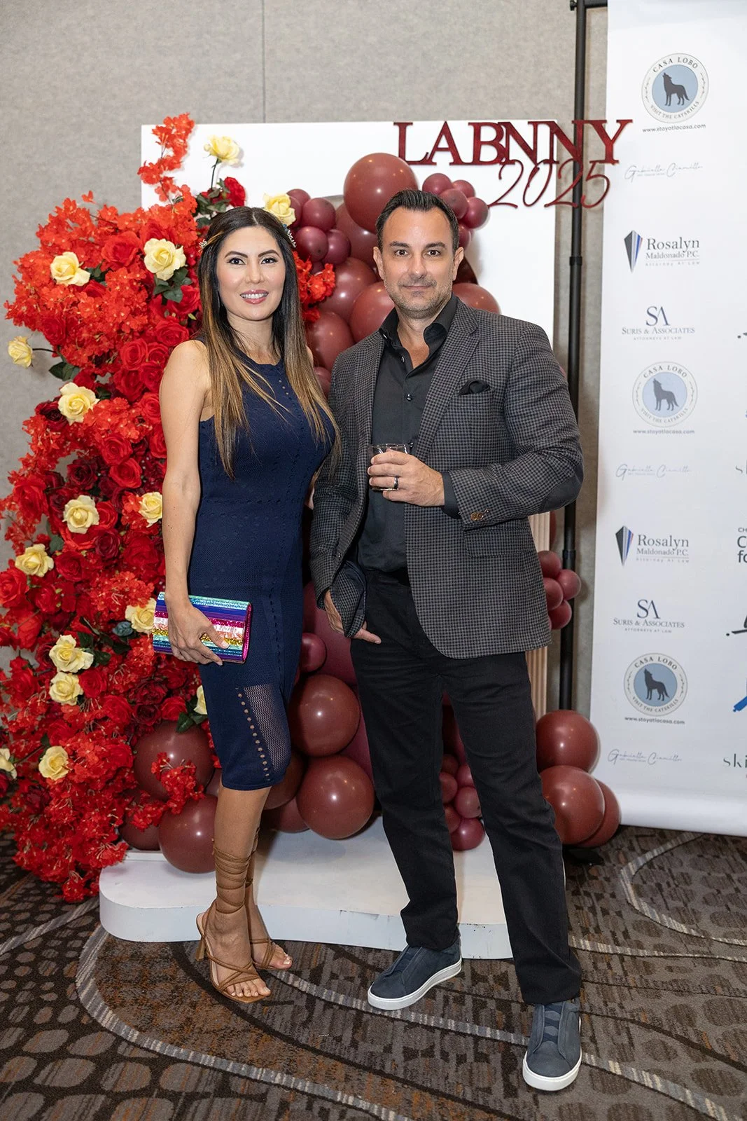 A woman in a navy blue dress and a man in a checkered blazer and black pants stand together at a formal event, in front of a decorative backdrop with red flowers, balloons, and a sign reading "LABN 2025".