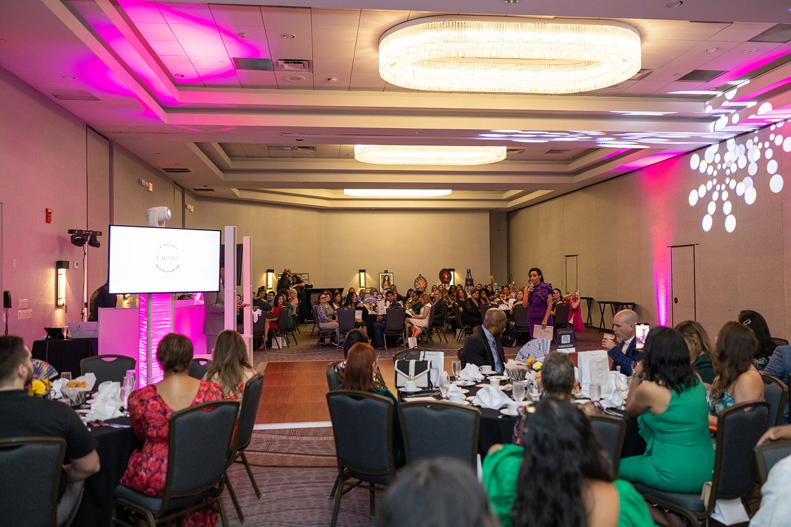 An indoor event with multiple round tables set for dining, decorated with flowers and tableware, with a woman in a purple dress speaking on stage, and a large screen, pink lighting, and decorations on the walls.