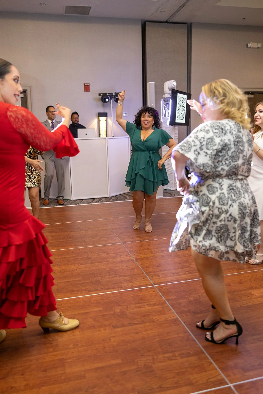 People dancing at a celebration or party, with women in colorful dresses and a DJ in the background.