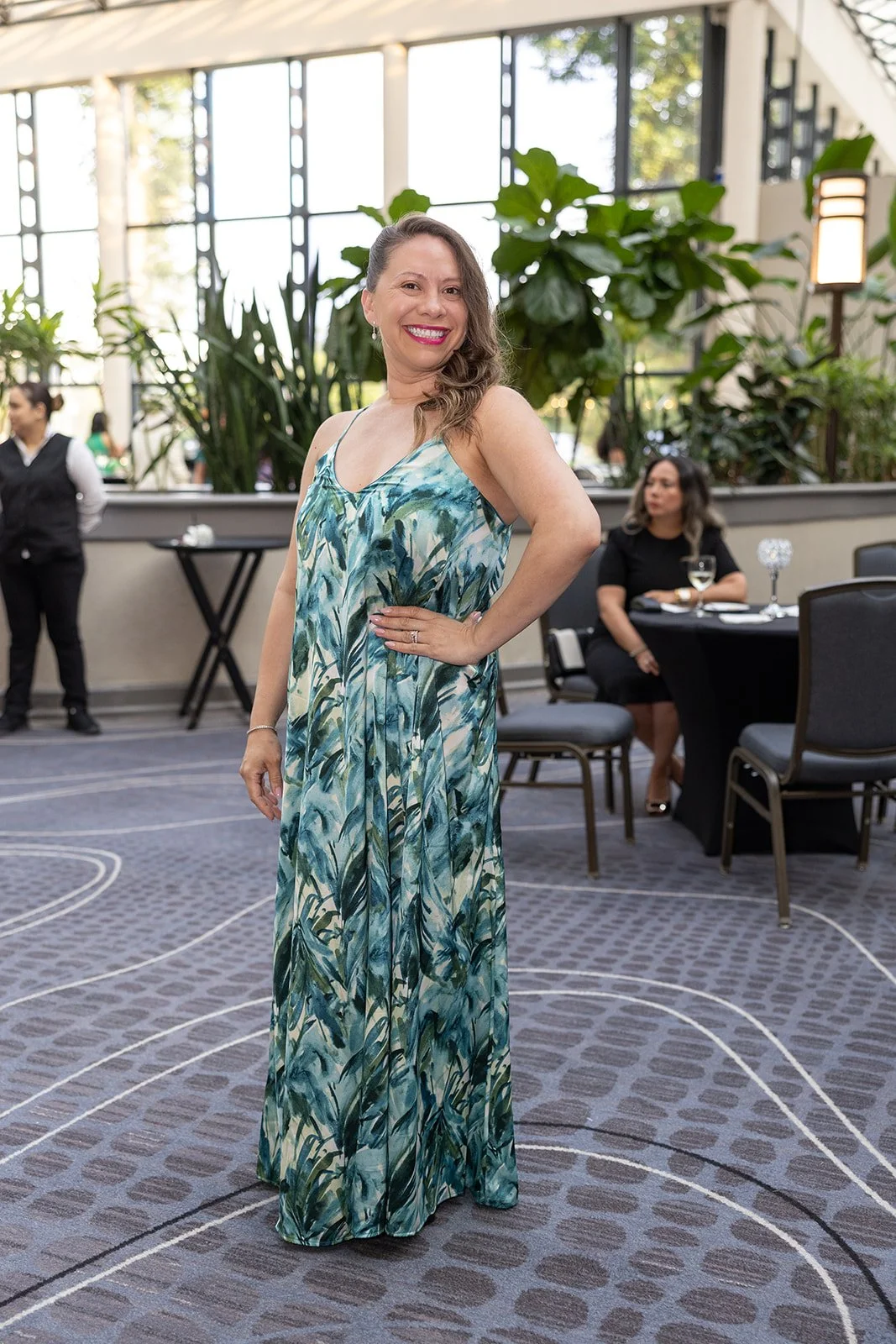 A woman in a colorful, tropical-patterned maxi dress smiling in a well-lit indoor space with large windows, plants, and other guests seated at tables.
