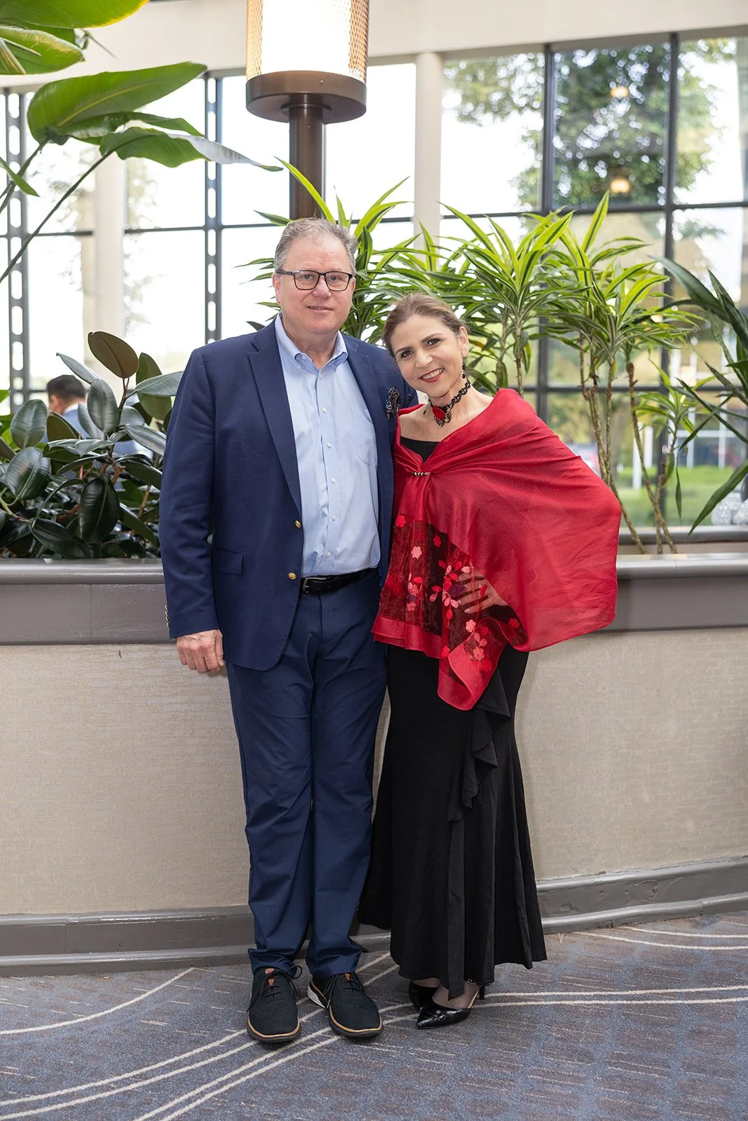 A man in a blue blazer and glasses standing next to a woman in a black dress with a red shawl, both smiling and posing indoors with large windows and green plants in the background.