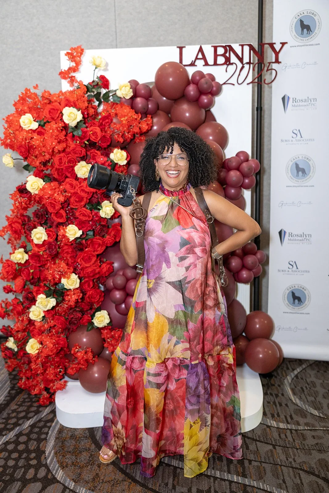 A woman with curly hair, glasses, and a floral dress holding a camera, standing in front of a backdrop with flowers and balloons at an event.