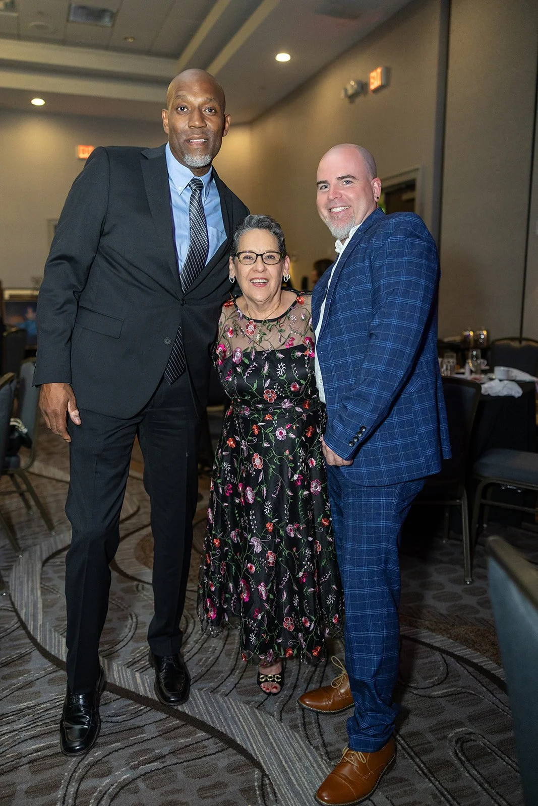 Three people standing together in a room, smiling at the camera. They are dressed in formal attire, likely at an event or gathering.