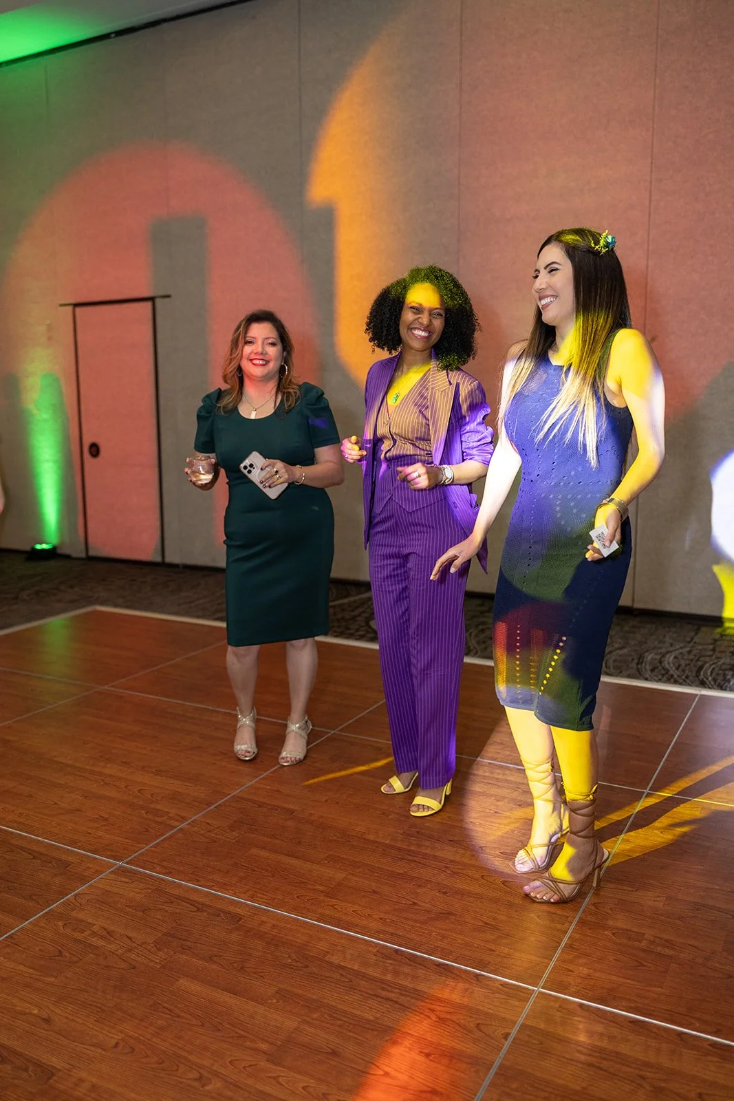 Three women are standing on a dance floor at an event or party, smiling and holding drinks. The background features colorful lighting.