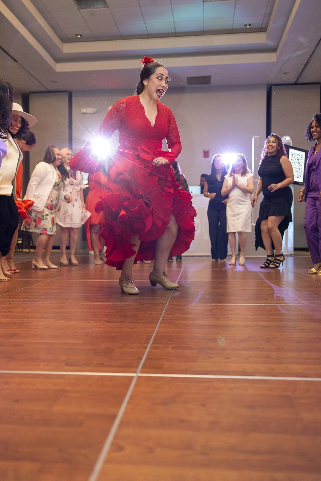 A woman in a red flamenco-style dress dancing on a wooden floor at a social event, surrounded by other guests watching and enjoying the dance.