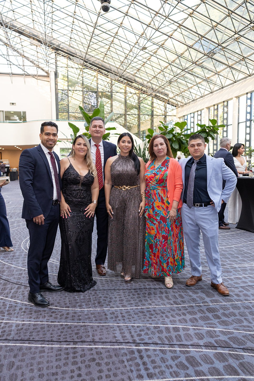 Group of six people posing for a photo at an indoor event, dressed in formal attire, with plants and a high glass ceiling in the background.