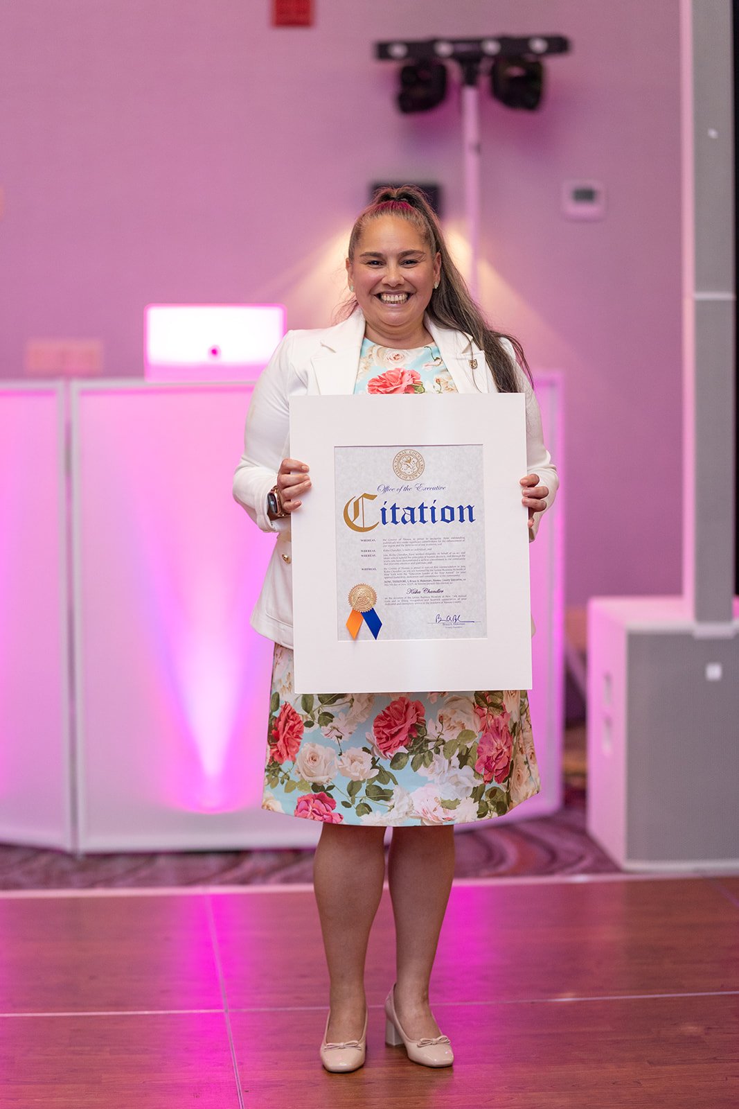 A woman with long hair tied in a ponytail, wearing a white blazer and floral dress, is smiling and holding a framed certificate.