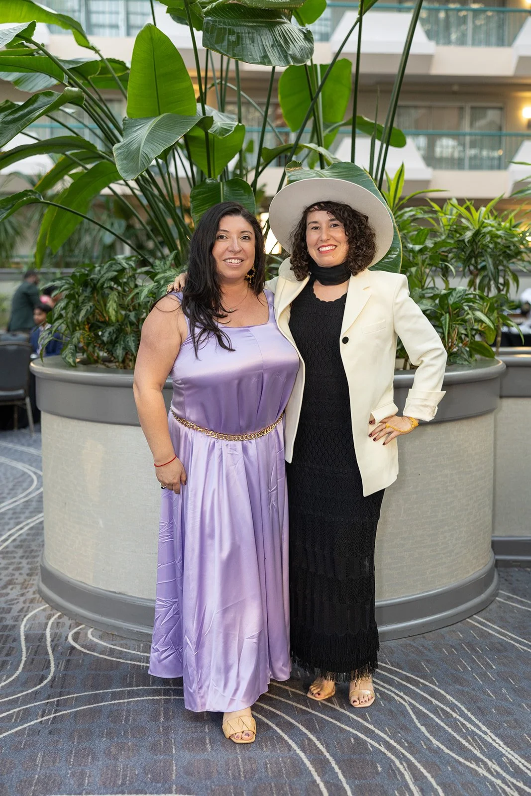 Two women standing close together in front of large indoor plants, smiling at the camera. One is wearing a lavender dress with a gold chain belt, and the other is dressed in a black dress with a white blazer and wide-brimmed hat.