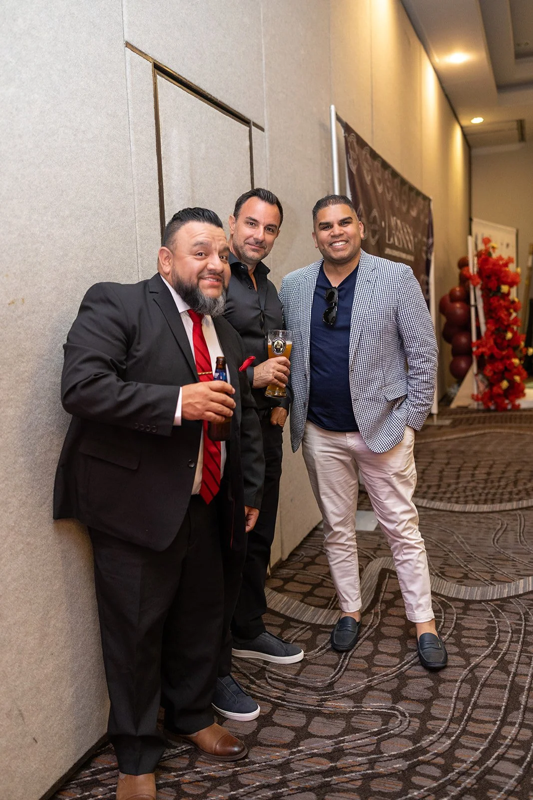 Three men standing together at a social event, holding drinks and smiling at the camera, in a hallway decorated with flowers and a banner.