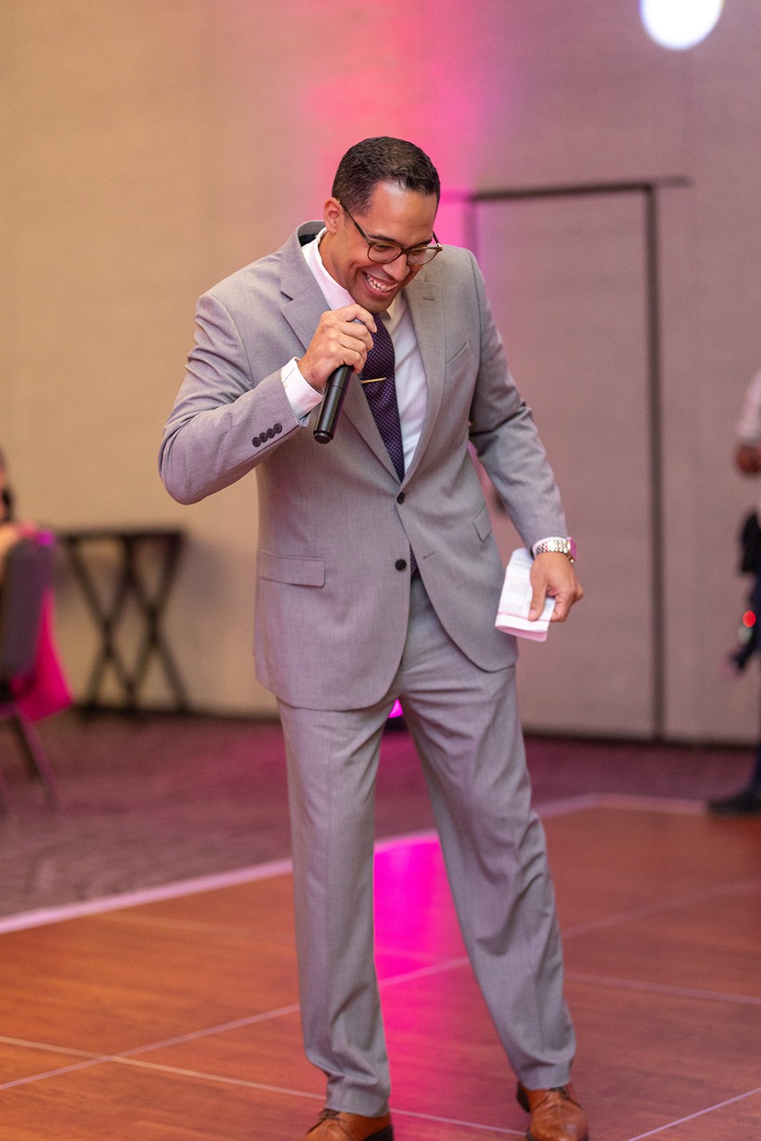 Man in a gray suit holding a microphone and smiling on a dance floor.