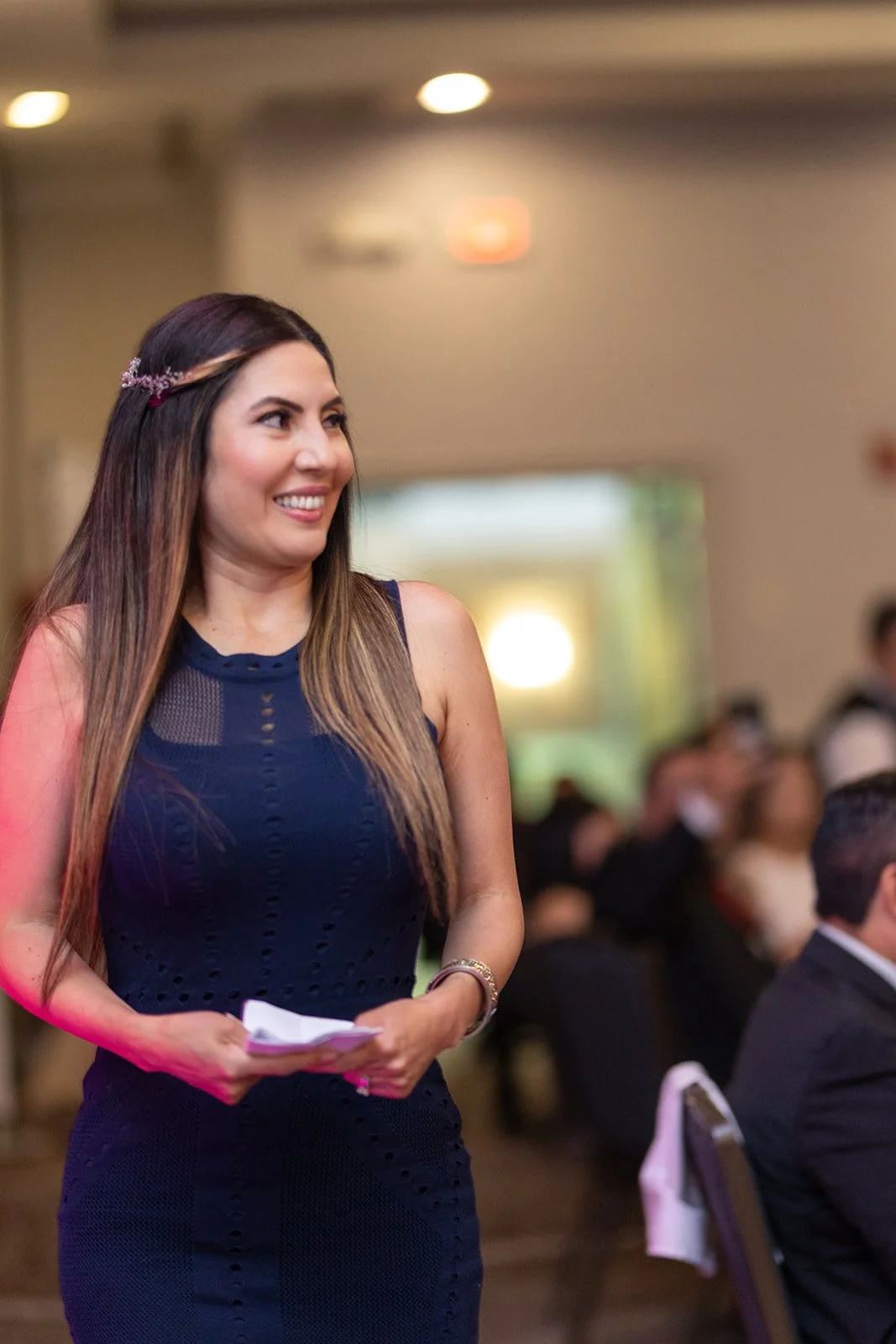 A woman in a navy blue dress standing and smiling at an indoor event with seated people in the background.