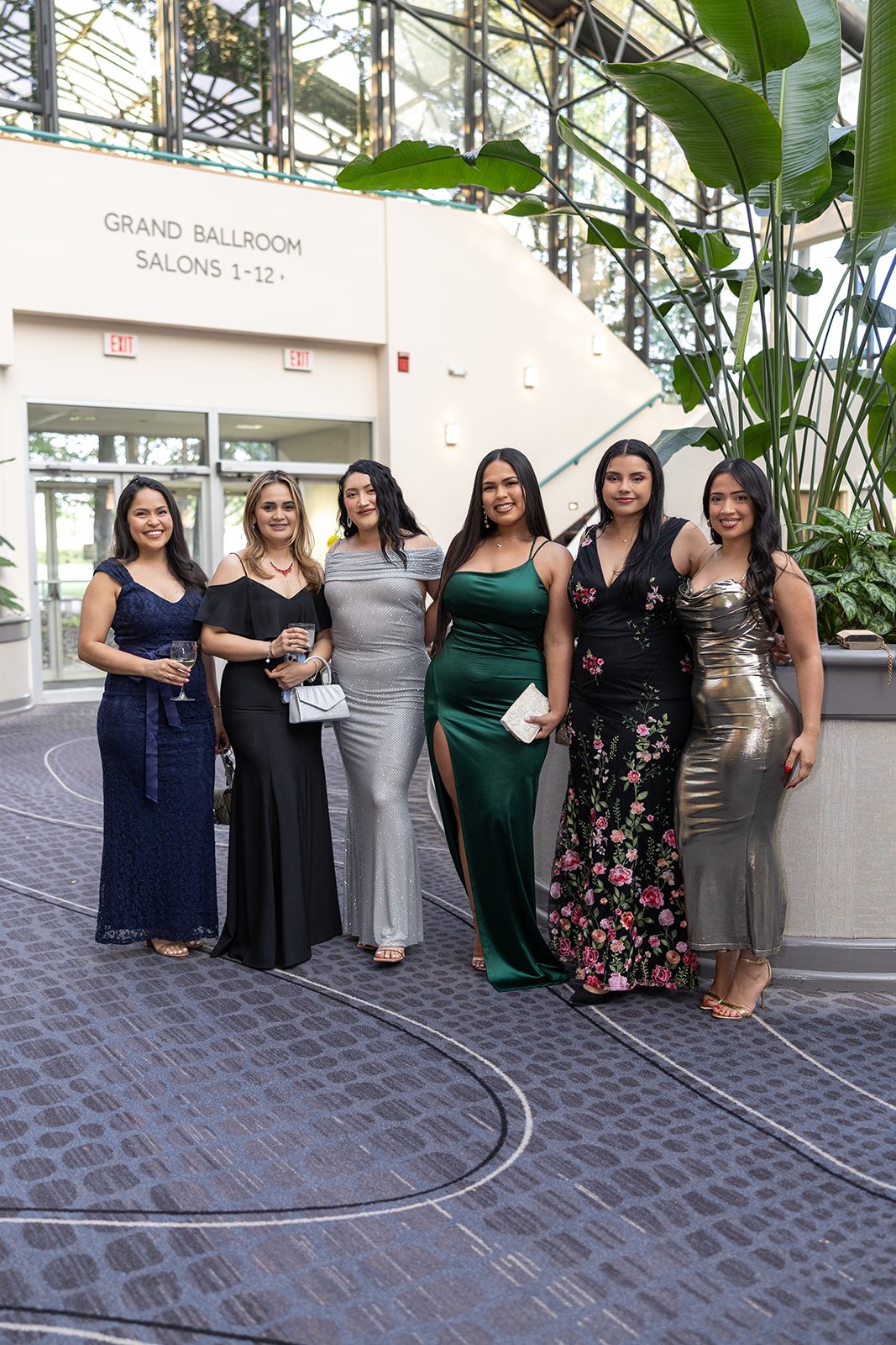 Six women dressed in formal gowns posing together indoors at a celebration or event.