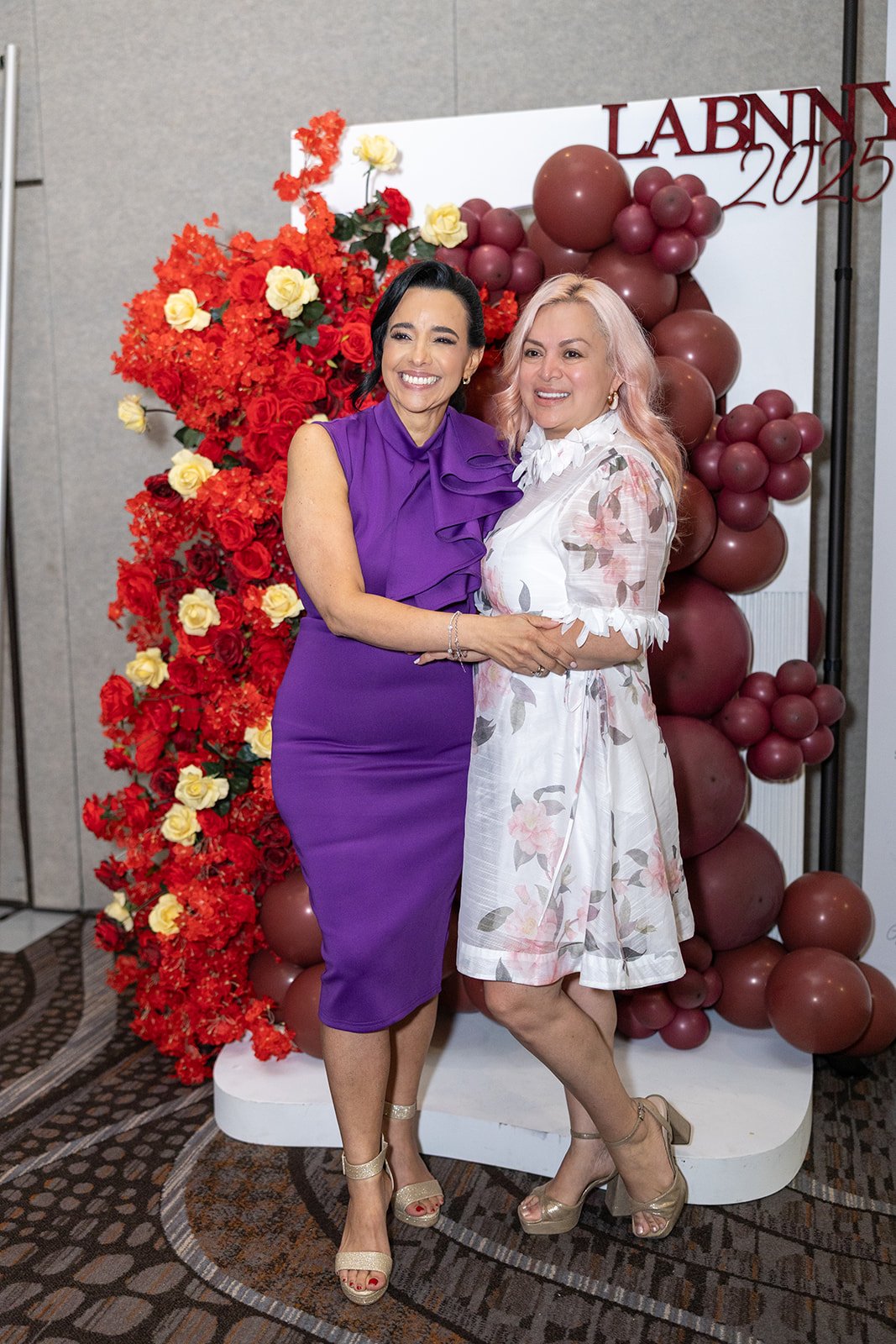 Two women standing together, smiling, in front of a flower and balloon backdrop at an event.