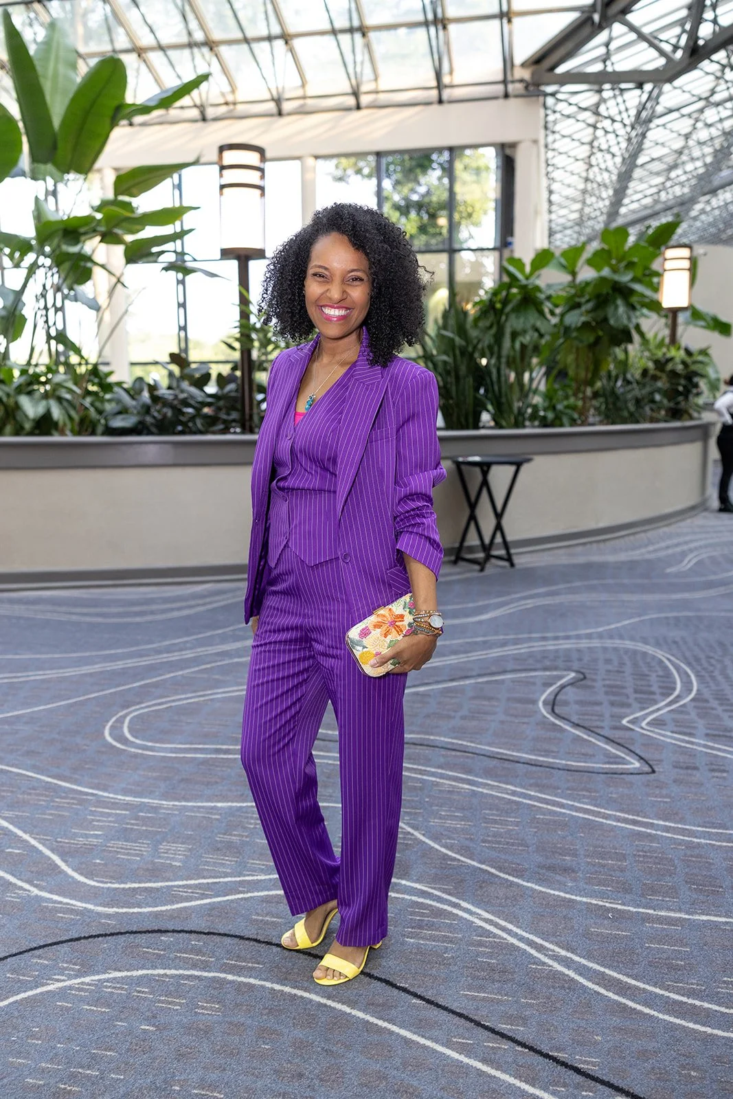 A woman in a purple pinstripe suit standing in an indoor space with large green plants, bright natural light, and modern lighting fixtures. She is smiling and holding a floral clutch purse.