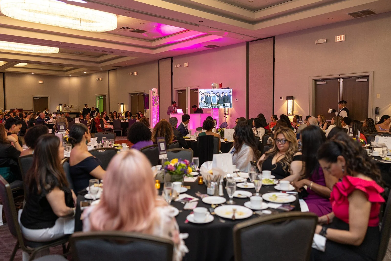 Guests seated at tables during an indoor banquet or fundraising event with a stage, large screen displaying a group photo, and pink lighting accents.